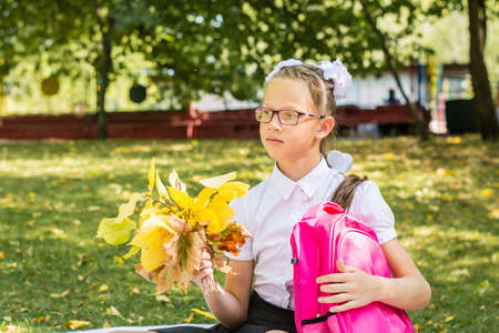 A cute schoolgirl with white bows is holding a bouquet of autumn leaves and a backpack in a sunny autumn park. Back to school concept. Copy spaceの写真素材