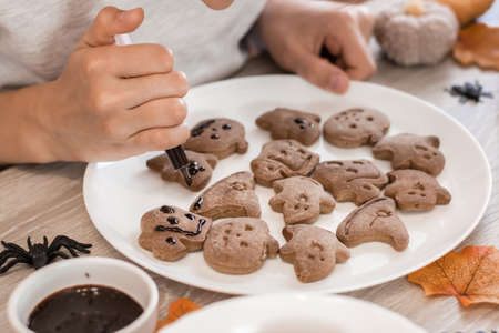 Children's hands squeeze chocolate icing from a syringe onto a halloween gingerbread cookie on a plate. Cooking treats for halloween celebration. Lifestyleの写真素材