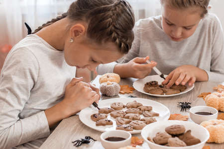 Two girls decorate halloween gingerbread cookies on plates with chocolate icing. Cooking treats for halloween celebration. Lifestyleの写真素材