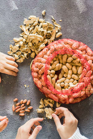 Mesh bag with inshell peanuts and two girls in the background are peeling nuts at the table. Lifestyle. Top and vertical viewの写真素材