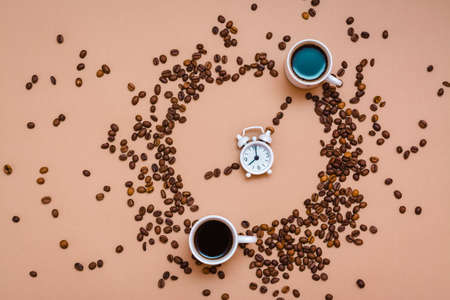 Two cups of black coffee in a circle from coffee beans and a white alarm clock on a beige background. Time to drink coffee concept. Top viewの写真素材