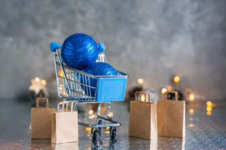 Holiday shopping. Shopping cart with blue shiny balls and paper bags on a gray background with garland and lights. Christmas conceptの写真素材