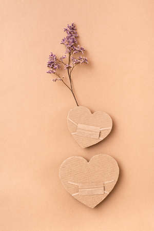 Pandemic date concept. Two masked cardboard hearts with dry flower together on a beige background. Top and vertical view. Valentine's dayの写真素材