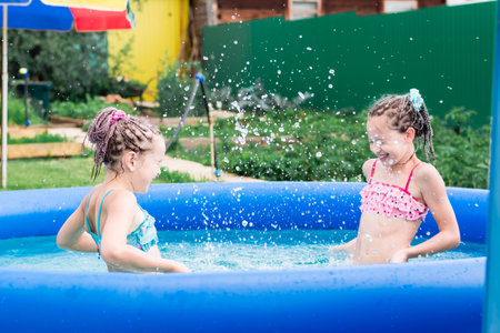 Two girls with afro-braids have fun splashing water in an inflatable pool on a summer day in the backyardの写真素材