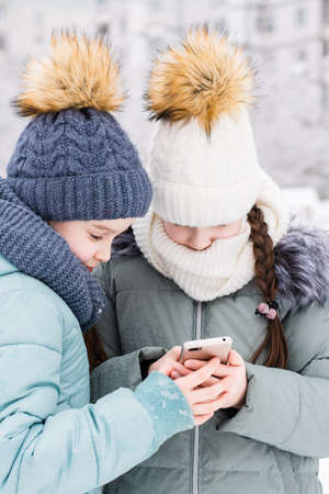 Two girls in warm coats and hats write a text message on a smartphone in a snowy winter park....