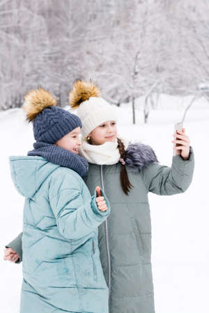 Two smiling girls in warm coats and hats take a selfie on a smartphone in a snowy winter park. Lifestyle use of technology. vertical viewの写真素材