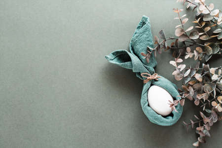 A white chicken egg is wrapped in a napkin in the form of a rabbit and a branch of eucalyptus on a green background. Eco friendly happy easter. top view.の写真素材