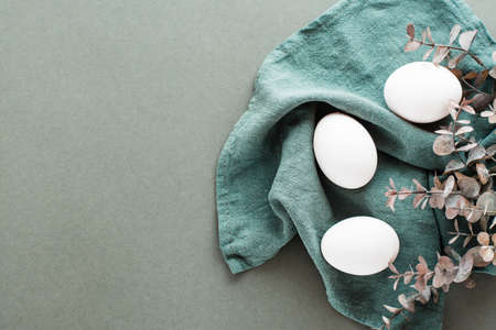 White chicken eggs on a linen napkin and a branch of eucalyptus on a green background. Eco friendly happy easter. top view.の写真素材