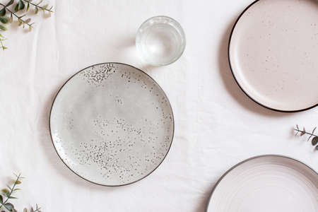 Empty ceramic plates, a glass of water and eucalyptus branches on a white cloth. top view. Eco...
