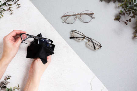 Women's hands clean glasses with a cloth. Glasses for vision in a metal frame on a two-tone background. Vision correction concept. top view.の写真素材