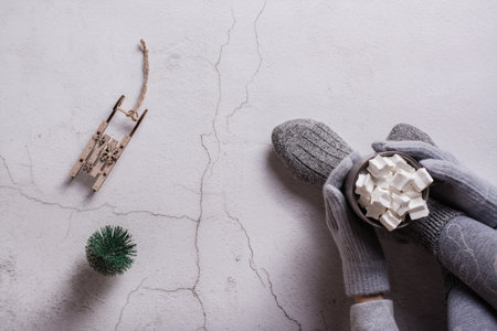 Women's hands in gloves hold cocoa with marshmallows, a Christmas tree and a wooden sledge. top view.の写真素材