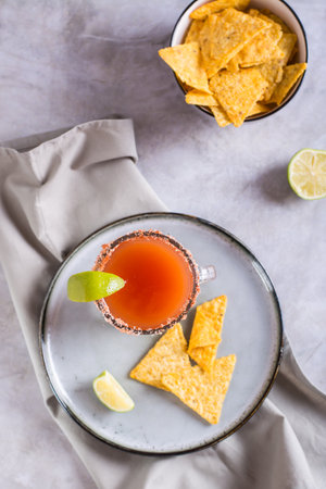 Michelagua mocktail with tomato juice and lime juice in a glass and nachos in a bowl top and...