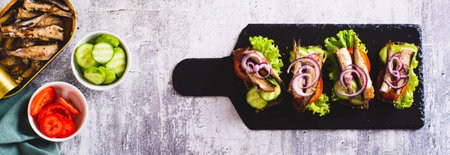 Sandwiches on rye bread with smoked sprats, vegetables and herbs on slate top view web banner