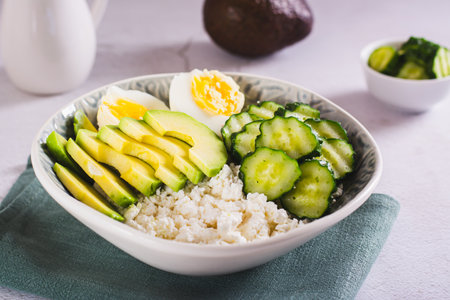 Bowl with cottage cheese, cucumber and avocado slices and boiled egg on the tableの写真素材