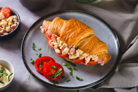 Delicious croissant sandwich with scrambled eggs, sausages and tomatoes on a plateの写真素材