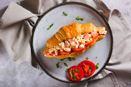 Close up of croissant sandwich with scrambled eggs, sausages, tomatoes and herbs on a plate top viewの写真素材