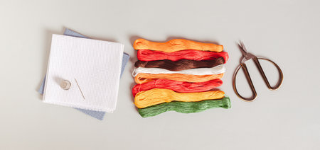 Set of colored floss, canvas and scissors for embroidery on a gray background top view web banner