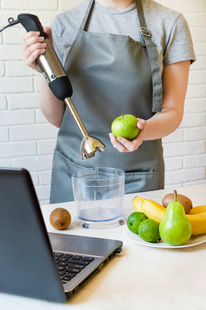 girl in an apron holds a mixer and an apple. making fruit smoothies according to a recipe from the...