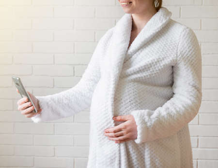 pregnant girl in a soft robe uses a smartphone to communicate.の写真素材