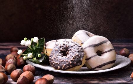 Donuts with chocolate and vanilla frosting on a brown platter, with hazelnuts and white flower decoration, sprinkled with powdered sugarの写真素材