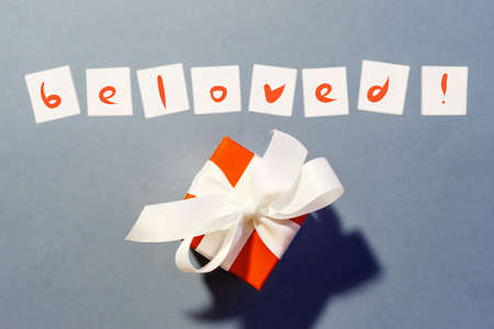 Ready lettering "beloved" on white squares of paper on a blue background and a box with a gift tied with a white ribbon.の写真素材