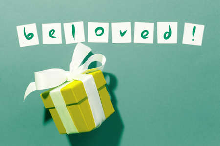 Ready lettering "beloved" on white squares of paper on a blue background and a box with a gift tied with a white ribbon.の写真素材