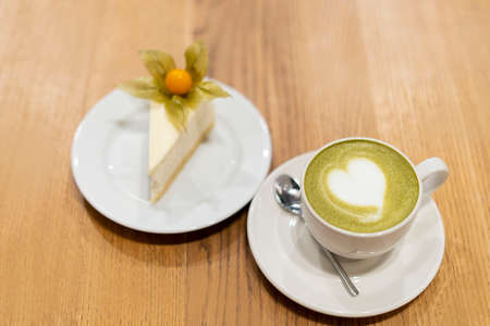 On the wooden surface stands a white cup of matcha tea. On a saucer is a white cheesecake with a...