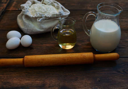 On the wooden table is a vintage jug with fresh milk, eggs, wooden rolling pin dough, olive oilの写真素材