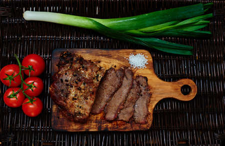 On the table lies a rattan board which served pork tenderloin baked in the oven in one piece, next leeks and cherry tomatoes from which cook fresh light saladの写真素材