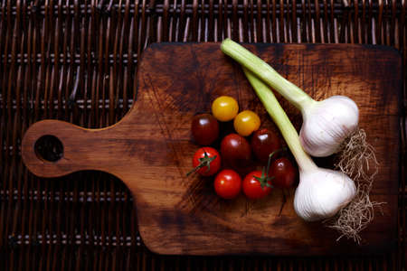 Two unpeeled garlic cloves and colorful cherry tomatoes are on vintage wooden cutting board,の写真素材