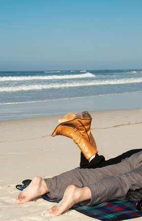 Couple lying on the sandy beach on holiday in winter. Woman is wearing black pants with brown leather boots and the man is wearing grey linen pants. He's got dirty feet from sand.の写真素材