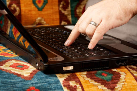 Close-up of Caucasian man's hand on black laptop keyboard. The man is sitting on the couch at home...