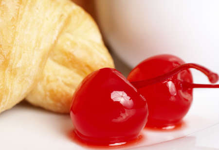 Macro shot of glazed maraschino cherries on white plate with fresh croissant at the backの写真素材