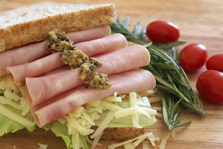 Tasty open sandwich with green lettuce, grated cheese, smoked ham and wholegrain mustard on wholewheat bread with rosemary and tomatoes on chopping boardの写真素材