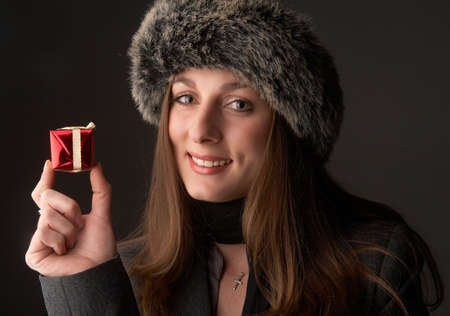 Portrait of a beautiful young brunette woman wearing a winter jacket and fur hat holding red gift box on black backgroundの写真素材