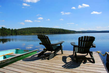 Two adirondack wooden chairs on dock facing a blue lake with clouds reflectionsの写真素材