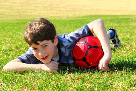 Young cute happy boy lying on grass with red soccer ball の写真素材