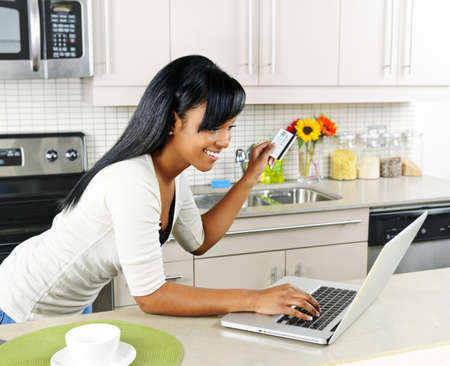 Smiling black woman online shopping using computer and credit card in kitchenの写真素材