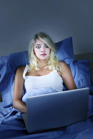 Scared woman using laptop computer in bed at night with wide eyesの写真素材