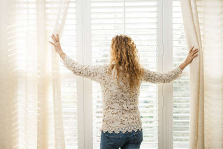 Woman looking out big bright window with curtains and blindsの写真素材