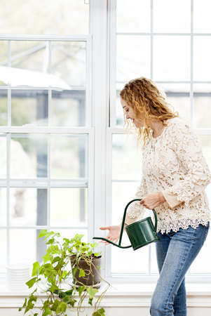 Smiling woman watering green plant at home by windowの写真素材