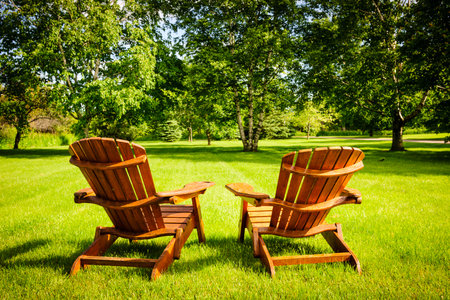 Two wooden adirondack chairs on lush green lawn with trees Two wooden adirondack chairs on lush green lawn with trees