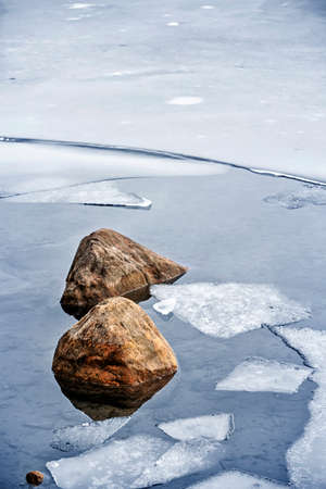 Broken ice floating on water at cold lake shore with rocks in winterの写真素材