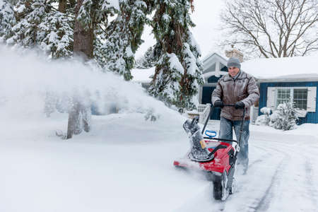 Man using snowblower to clear deep snow on driveway near residential house after heavy snowfall.の写真素材