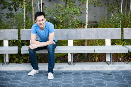 Young asian man sitting and smiling on park bench with copy space