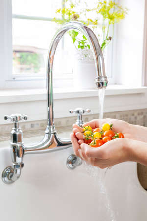 Hands washing fresh cherry tomatoes in running water of kitchen sink with curved faucetの写真素材