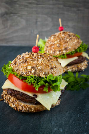 Two gourmet hamburgers with swiss cheese and fresh vegetables on multigrain buns over dark backgroundの写真素材