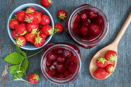 Glass jars with homemade strawberry preserve and fresh strawberries in bowl from above on blue rustic wood backgroundの写真素材