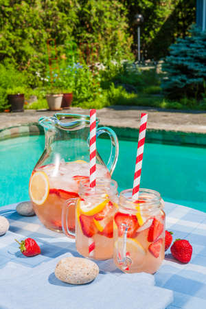 Ice cold homemade strawberry lemonade in jug and glasses with paper straws on outdoor pool side table in summer with garden in backgroundの写真素材
