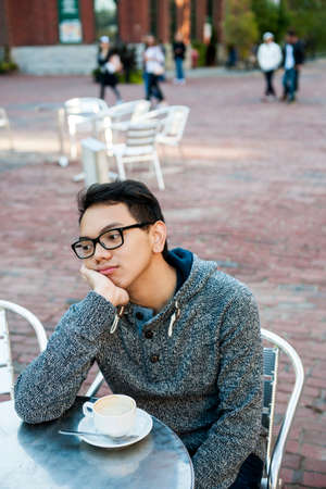 Young asian man in outdoor cafe with cup of coffee looking away being bored and unhappy